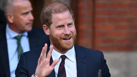 Prince Harry wearing a suit, waving towards a camera.