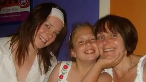 Family photo A teenage girl (left) with brown hair in a white headband poses with a younger sister with brown hair, who is wearing a white flowery dress and her mother, who has brown bobbed hair.