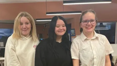 Shyanne, Libby and Shanice stand side by side. Shyanne has blonde hair and wears a white hoodie. Libby has black hair and is wearing a black jumper. Shanice has her hair tied back, wears glasses and a white shirt with short sleeves. 