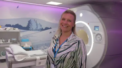 Jamie Niblock/BBC Claire Moore with long hair. She has a light coloured top on and a blue lanyard. She is standing in front of a MRI machine.