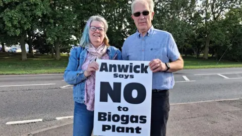 LDRS A man and woman holding up a white and black sign which reads 'Anwick says no to biogas plant'. The woman is wearing double denim and has colourful hair and glasses and the man is wearing black sunglasses and a blue shirt.