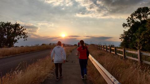 MONDAY - Two women walk away from the camera, one is in a long sleeve white top, the other is in a red t-shirt. They both wear black tracksuit bottoms and trainers. The sun is setting on the horizon over a motorway bridge. To the left of the picture is a road and on either side the verges are dry yellowing long grass.