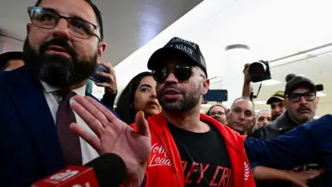 Reuters Tarrio in sunglasses and a red sweater gestures towards a microphone in the middle of a crowd of people.