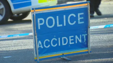 BBC A sign with 'POLICE ACCIDENT' in bold white letters on a blue background on a road, with a police vehicle behind it.