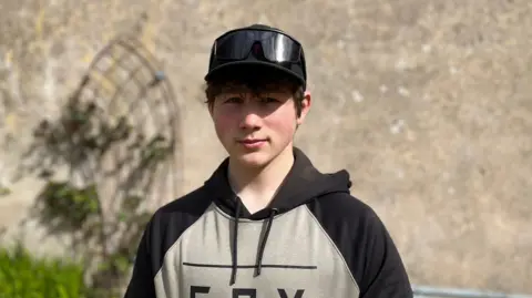 A boy with short dark hair, wearing a black baseball cap with sunglasses placed on top. He is also wearing a grey and black hooded jumper. He is staring directly at the camera.