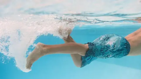 A young boy swimming under water in a pool, he has blue swim shorts on and is kicking in the water.