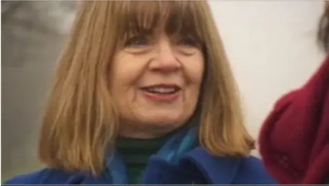 Close-up of Paula Hiscocks with shoulder-length hair wearing a blue coat and blue scarf outdoors. The image is softly focused, with a blurred background and part of another person in a red coat visible at the right edge of the frame.