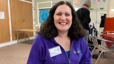 BBC Foodbank Manager Emma Murray wearing a purple foodbank zip up fleece with shoulder length dark hair smiling at the camera in a hall 