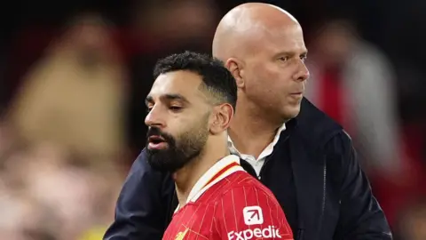 PA Media Mohamed Salah looks unimpressed as he leaves the pitch after a recent game and is embraced by Arne Slot, who is pursing his lips. Salah is wearing his red LFC strip while Slot wears a dark coat.