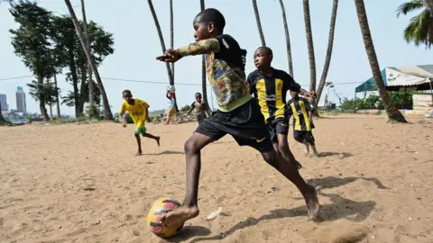 Issouf Sanogo / AFP Barefoot boys in football kits play football on a sandy pitch in Abidjan. Palm trees can be seen in the background - Wednesday 26 February 2025.