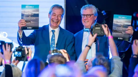 PA Media Nigel Farage and Malcolm Offord smile as they hold up Reform party manifestos with journalists holding up cameras in front of them, in Holyroo on 19 March.
