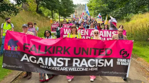 Pro-immigration demonstrators at the beginning of the march on Saturday. There are dozens of people with a variety of banners. The one at the front is a pink and black Stand up to Racism banner and says Falkirk United Against Racism. refugees are welcome here.