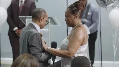 Yvonne Johnson Orville (left) in a suit and Yvonne (right) in a silver dress smiling at Orville holding a microphone. The pair are stood at their renewal ceremony in front of balloons and their friends and family.