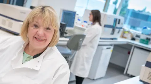 Cancer Research UK Sharon Quennell with blond bobbed hair stands in Cancer Research UK Manchester Institute's research lab wearing white coat. She is smiling. A researcher also in a white lab coat stands behind her.