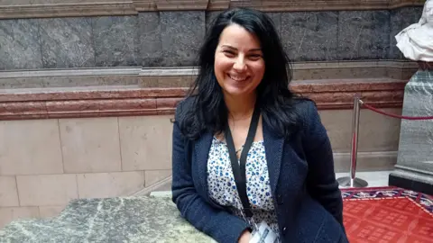BBC/Lucy Ashton A woman with long black hair is wearing a navy jacket and blue floral top. She is standing on a marble staircase in Sheffield town hall and smiling