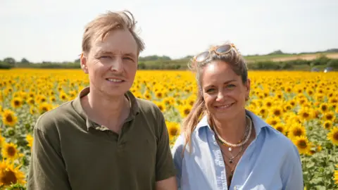 Shaun Whitmore/BBC Michael and Sophie Landale, tenant farmers at Westgate Farm in Walsingham. They are both looking directly at the camera and are smiling. They are both standing among their 30-acre sunflower field. Michael is on the left and is wearing an olive green short-sleeved polo shirt. Sophie, on the right, is wearing a pale blue shirt, with a number of necklaces round her neck and has a pair of sunglasses rested on her head.