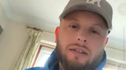 Family Ryan Griffiths wearing a grey cap and a blue hoodie. He has a short brown beard and moustache