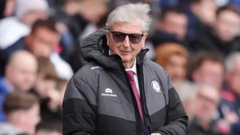 PA Media Football manager Roy Hodgson is pictured standing on the side of a pitch wearing a Bristol City black coat, a white shirt and burgundy tie and a pair of black sunglasses. He is looking to the side and appears to be smirking. The crowd behind him is blurred out.