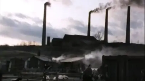 BBC/YouTube A slightly blurry image of four industrial chimneys with smoke pouring out, next to a large building. In the foreground is a film set of a Victorian village.
