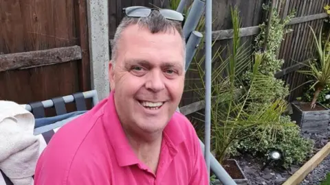 Family handout A man in a pink polo shirt smiles as he sits on a swinging chair in a garden. His glasses are pushed back on his head. His hair is brown and greying at the sides.