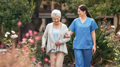 shapecharge/Getty Images A nurse and a woman with a walking stick in an outdoor space