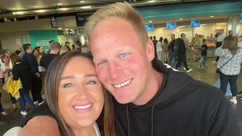 A couple take a selfie photo - on the right is a man with short blonde hair and blue eyes wearing a black hooded sweatshirt. He has this arm around a shorter woman with long brown hair wearing a white top. They are both smiling at the camera.