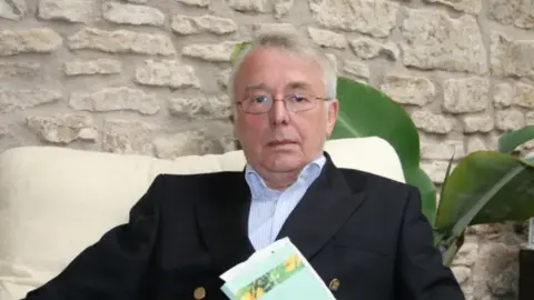 David Raynes David is sitting on a white chair with a large brick wall behind him. 