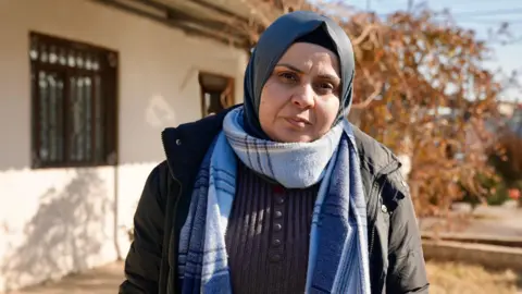 Goktay Koraltan/BBC Hekmiya Ibrahim stares into the camera. She is in a grey headscarf and a black puffer jacket, wearing also a light blue striped scarf for warmth