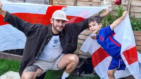 Jessica Beames Daniel Beames, wearing an England shirt and dark shirt, and his partner's son, Barney, wearing a blue England shirt, both hold England flags aloft in a garden in Bishop's Cleeve during the Euros in 2021.