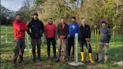 Katie Reynolds A group of people stood on a green field with trees behind them. Three of the people have spades and tools in their hands. All of the people in the photograph are smiling. 