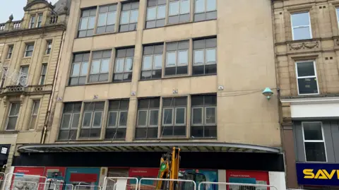 Photo shows a five-storey building on Fargate shopping centre. At the bottom of the building fences, bollards and a digger are blocking the entrance. The building has rows of windows on each floor but is in need of repair. It is not a historic building.