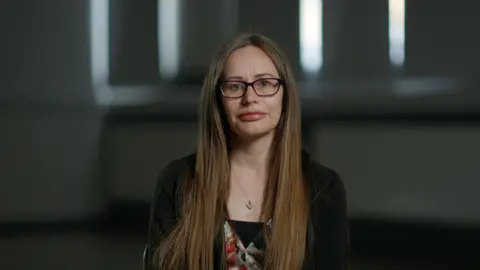 A photo of Susan Christie. She is looking at the camera against a dark internal background. She has long brown hair and glasses.