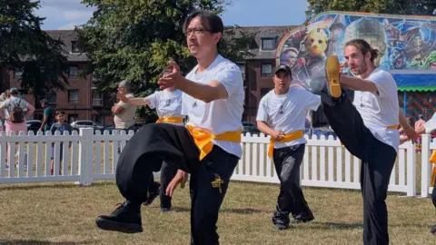 Art Asia A group of performers in white T-shirts and yellow belts perform kicks in front of a crowd. 