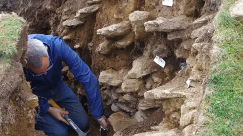 Historic England A man with glassed in blue overalls kneels in an archeological trench holding a hammer. Stones jut out from the side of the trench and a small white label is attached t one of them.