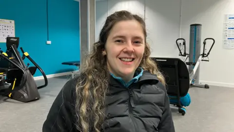 Jennifer is smiling at the camera. She has wavy blond hair and a blue fleece beneath a black waterproof coat. There are machines behind her.