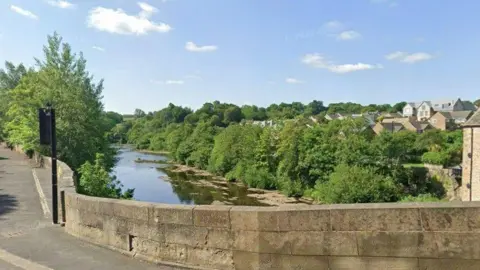 A view over County Bridge in Barnard Castle over the River Tees. Both sides of the river are covered in greenery and trees. A traffic light can be seen on the bridge to the left.