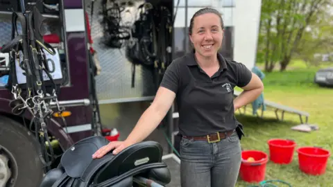Jade stands in front of a lorry which has tack hanging from hooks and a black saddle is on a stand to her right.