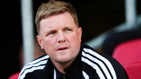PA Media Eddie Howe sitting down on a red chair during the Bristol game. He is looking to the right and has his mouth slightly open. He is wearing a black sports jacket with the white Adidas stripes on the shoulder.