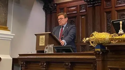 LDRS A man in a blue suit with a red tie and glasses, standing behind a wooden lectern in the city council building.