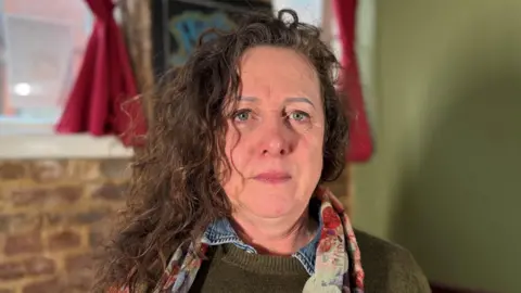 Ann Carver sits indoors, in front of a brick wall and a window framed with deep red curtains. A picture frame hangs on the wall in the background. Ann is wearing a knitted, dark olive-green top layered over a collared garment. Around her neck is a colourful scarf with a floral-like pattern in shades of red, cream, and muted blues. Her hair is long, wavy, and loosely draped over one shoulder.