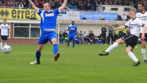 Hereford United Bowen, in lack and white kit, on the far right, has kicked the ball during a game with an opposition player, in blue kit, raising his arms out of the way as he raises his left leg. Crowds can be seen behind them in the ground and club member standing by the pitch.