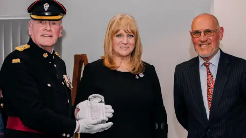 A man in military dress and white gloves shows a glass plague towards a camera. He is stood next to a woman with light hair in the centre and a man with a pinstriped suit on the right.