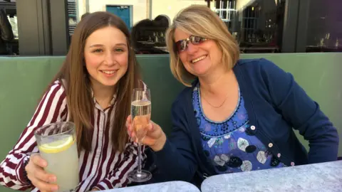 Ellie Gould wearing a white and burgandy striped shirt, holding up a glass of lemonade and smiling at the camera. She is sitting beside her mother, Carol Gould, wearing a blue patterned blouse and navy cardigan, holding up a glass of champagne.