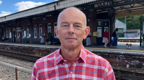 Nick Hurrell has thin, short, grey hair and he is wearing a red and white checked shirt as he stands in front a railway train station.
