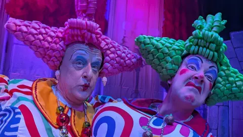 BBC Two panto dames are wearing brightly-coloured costumes and exaggerated make-up. The dame on the left is wearing a bright pink pointy plastic wig. The dame on the right is wearing green pointy plastic hair.
