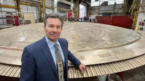 A person in a pinstripe suit and patterned tie stands inside an industrial workshop next to a large bronze structure with engraved lettering. The background shows yellow metal framework, machinery, and workers in red protective clothing.