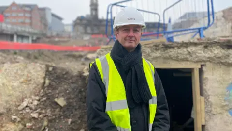 BBC/Simon Thake Martin Gorman, wearing a hard hat and bright yellow jacket, stands in front of stone remains and an open chamber