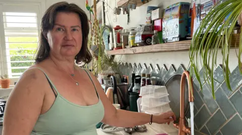 A lady standing in a kitchen next to a sink. The person is wearing a light green tank top and with one hand turning on a tap. The kitchen has various items on the countertop, including jars, bottles, and containers. There are also plants hanging above the sink and placed on the windowsill. The backsplash behind the sink features blue-grey diamond-shaped tiles.