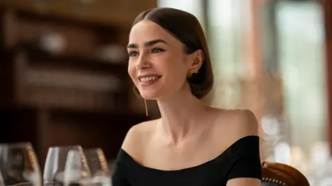 A production still image of Lily Collins as Emily, pictured smiling in a restaurant. She has short brown bobbed air, and wears an off-the-shoulder black top and gold earrings.