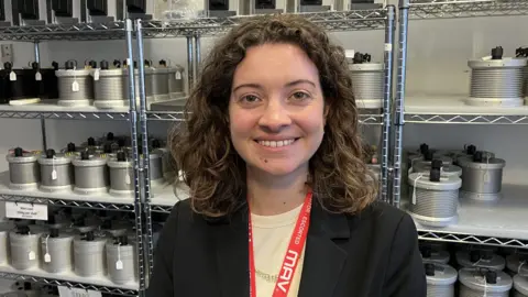 Sarah Coombes smiling for the camera, wearing a dark blazer. Shelves of metal camera units are visible behind her.